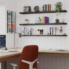 A home office corner featuring three sleek dark wood floating shelves with invisible brackets filled with books, plants, and desk accessories—perfectly complementing the clean-lined workspace beneath.