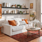 Two 72 inch long floating shelves in a ebony finish with invisible brackets styled with books and vases over a cozy white couch. Warm tones and framed art create a welcoming living room aesthetic.