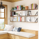 Sunlit nook with two wall mounted bookshelves displaying books and green decor pieces, adding warmth and charm to the space beside a window seat.