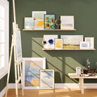 An artist's corner with a green wall displaying colorful paintings on natural wood floating shelves, a canvas on an easel, and a table with painting supplies.