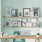 A workspace with a laptop on a wooden desk, a teal chair, and shelves for wall storage above holding various framed architectural drawings and photographs.