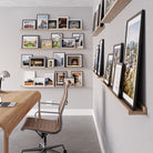 Modern office with a wooden desk and chair, set against a corner adorned with long floating shelves full of framed photos and sketches.