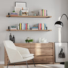 Two wood floating shelves with black brackets, styled with books, candles, and decor items. Paired with a light wood dresser, white armchair, and sleek black lamp, creating a modern, cozy, and minimalist vibe.
