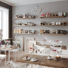 A cozy coffee shop with hanging book shelves holding coffee products, mugs, and decor. White chairs and wooden tables are decorated with pink flowers and pastries, creating a warm, inviting ambiance with natural light.