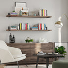 A cozy living room featuring two wooden floating shelves in rich espresso custom finish holding books and decor. Below, a wooden dresser supports vases and candles. A white chair, gold floor lamp, and black coffee table complete the stylish setup.