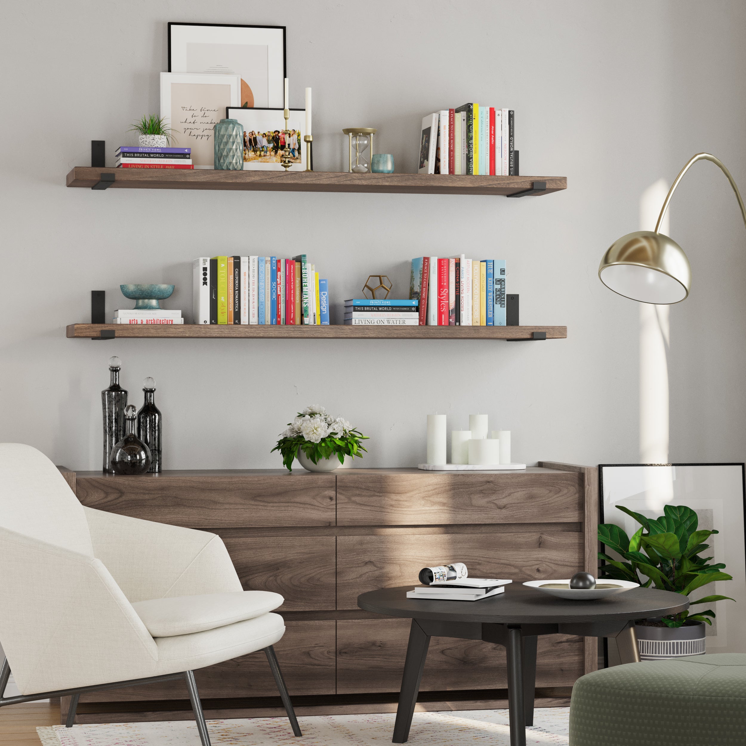 A cozy living room featuring two wooden floating shelves in rich espresso custom finish holding books and decor. Below, a wooden dresser supports vases and candles. A white chair, gold floor lamp, and black coffee table complete the stylish setup.