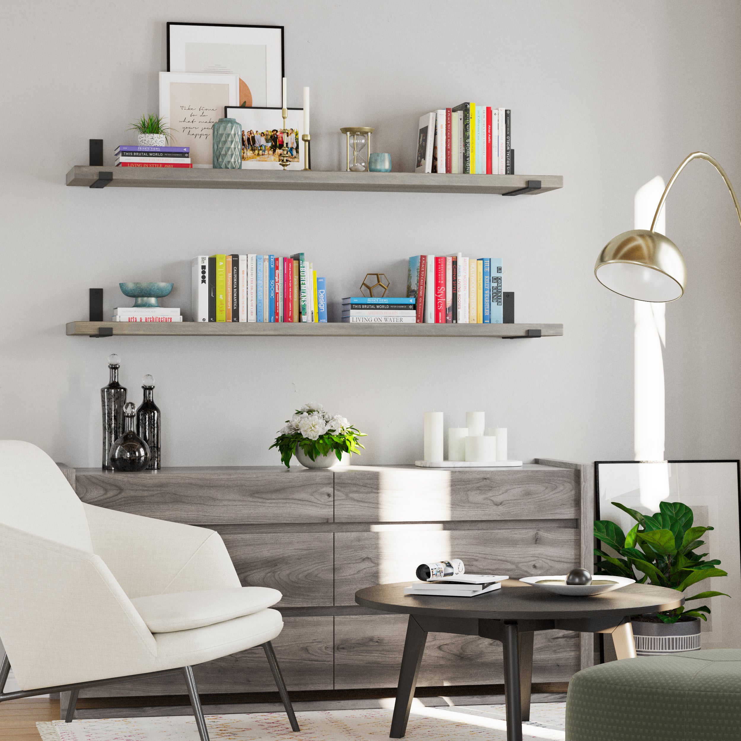 A cozy living space with gray floating bookshelves displaying books, decor, and plants. Below, a wooden dresser holds candles and flowers. A white chair, round coffee table, and gold lamp complete the stylish arrangement.