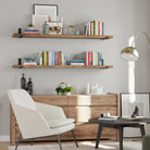 A cozy reading corner featuring two long display shelves in rich provincial finish with books and decor above a wooden dresser. A white armchair, black round coffee table, and gold floor lamp complete the modern, elegant design.