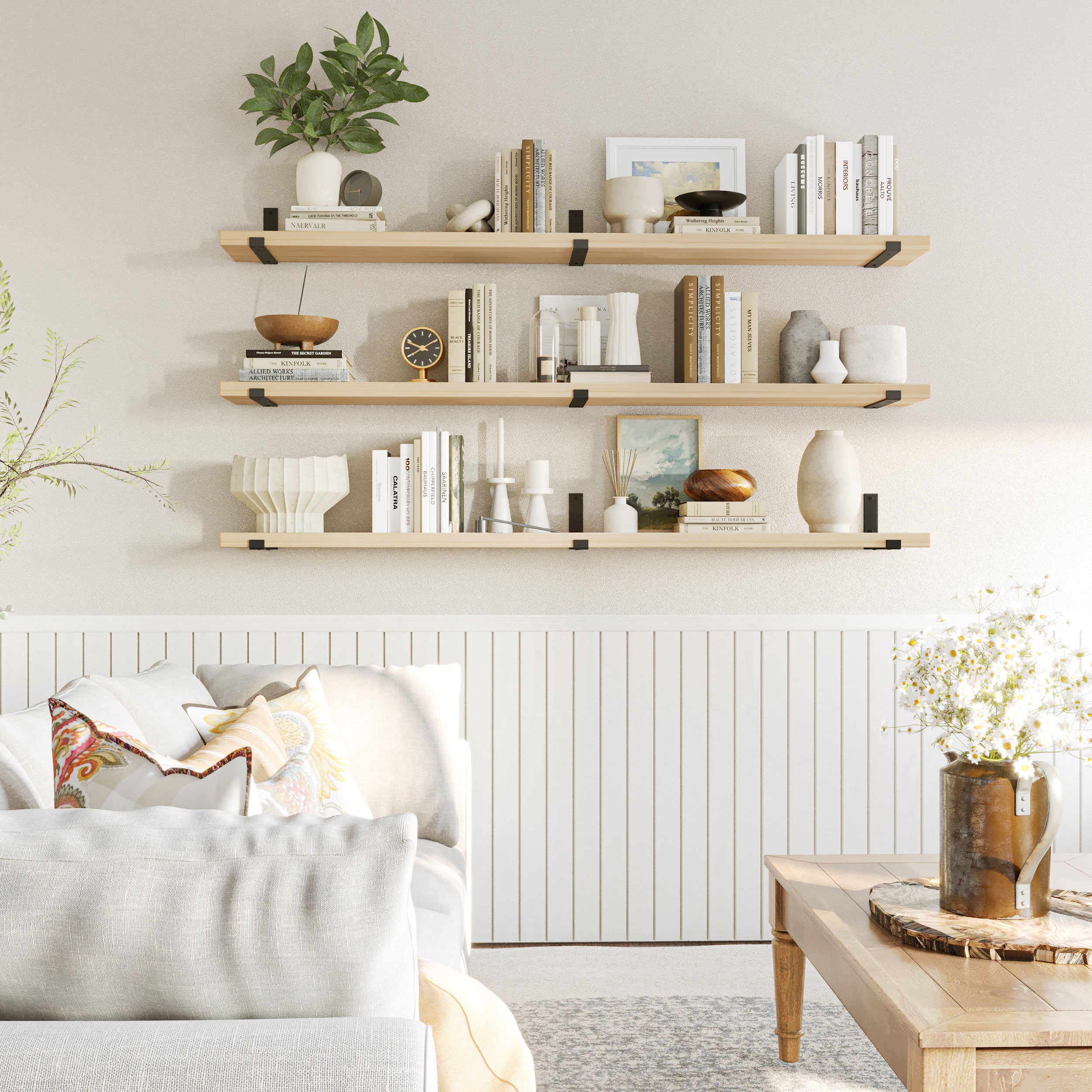 Three floating shelves in Natural finish hold books and ceramics above a white wainscoted wall, complementing a cozy and bright living room with neutral tones.
