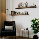 Two dark walnut real wood shelves styled in a cozy living room above a chair. Decorated with books, plants, and frames, showcasing modern wall storage and decorative functionality.