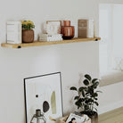 Golden oak wall mounted shelf mounted above framed art and plant. Decorated with books and vases, creating a cozy, modern wall display in a bright living space.