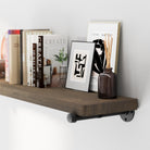 Wooden shelf with pipe brackets with books, a framed picture, and a vase against a white wall.