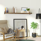 A single rustic wood wall shelf sits above a small wood side table with a cozy reading chair. Books, décor, and framed photos add a stylish accent to the corner, warmed by soft lighting and neutral tones.