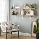 Bright living space featuring a two-tier adjustable wall shelf for living room decor with light wood boards and white brackets, styled with books, ceramics, and greenery above a cozy chair.