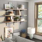 Styled living room decor featuring a 3-tier shelf with black brackets and wood boards, decorated with framed art, vases, books, and plants near a window.
