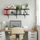 Styled home office scene with black floating 2 tier wall shelf mounted above a desk, decorated with books, framed art, plants, and modern accessories.