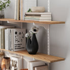 Wooden shelving unit with white metal bracket, featuring stacked books and a black vase with flowers, highlighting wood grain detail and sturdy bracket support.